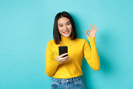 电子商业和网上购物概念 亚洲女性的肖像显示OK标志和使用手机 赞美App 站在蓝背景上站立青少年交易发型学生手势女孩黑发理发促销图片