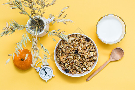 健康健康的早餐 碗中的烤面包 牛奶杯 橙子 木勺和黄色背景的闹钟 最高风景图片