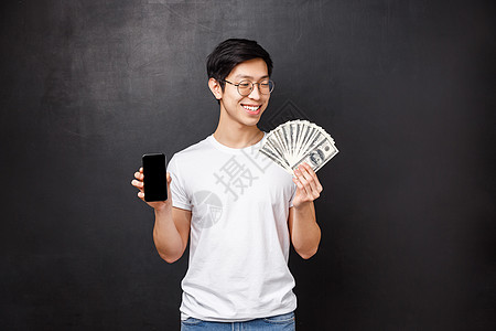 技术 金钱和奖品概念 身穿 T 恤 面带微笑 快乐的亚洲富豪 对现金的美元粉丝感到满意 手持手机 展示智能手机屏幕 黑色背景图片