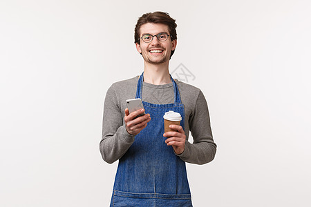 快乐的年轻男性员工的肖像建议使用带有手机应用程序的促销代码在他的咖啡店获得折扣 拿着外卖杯和智能手机微笑相机图片