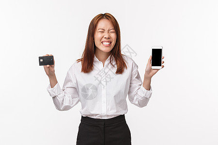 信用卡中兴奋 快乐 微笑的亚洲女性的肖像 展示手机显示屏 闭上眼睛 笑着最终点了她想要的东西 善待自己 互联网和金融概念图片