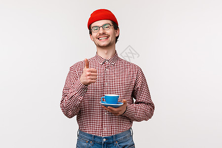 戴着眼镜和红色豆豆的留着胡须的男人高兴而轻松地笑着 竖起大拇指 拿着茶或咖啡的杯子 对制作精良的卡布奇诺咖啡感到满意 站立的白色图片