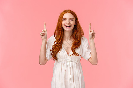 快乐幸运的红发欧洲女性穿着时尚的白色连衣裙 指着手指对着镜头笑 广告宣传 展示公司横幅 最喜欢的美容产品图片