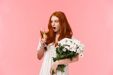 女孩对香槟的味道印象深刻并感到惊讶 迷人的红发女性庆祝 浪漫约会 持有白色花束和一杯饮料 粉红色背景图片