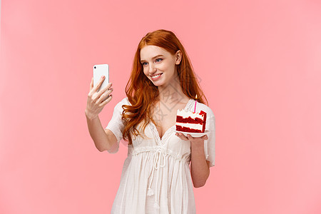 穿着白裙子红长头发的caucasian寿终女孩 在每天晚宴期间自拍在网上社交媒体上张贴 手持智能手机和和平美味蛋糕 粉红色背景图片