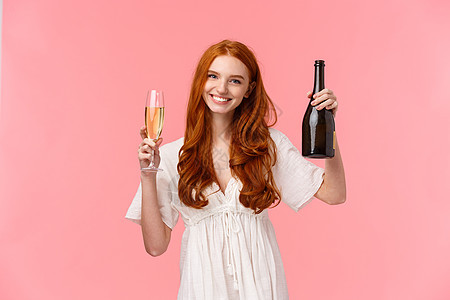 干杯 恭喜我的朋友 身着白裙 举起酒杯 举着香槟酒瓶 庆祝成功 粉红色背景的温柔 快乐 长相友善的迷人红发女性图片