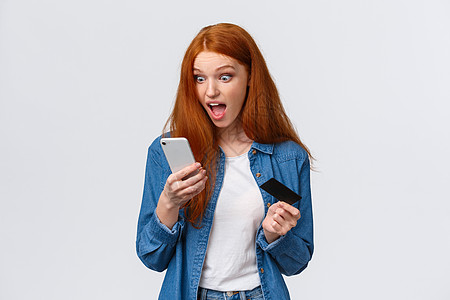 兴奋和震惊的年轻红头女红发女孩匆忙买裙子 打折扣超棒 盯着印象深刻 令人惊讶的智能手机显示器 持有移动电话和信用卡以及白背景图片