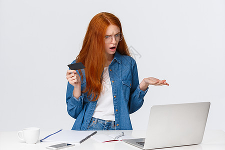 困惑和困惑的红发女人无法进行网上购物 不知道为什么会出现转账问题 沮丧地耸耸肩指着笔记本电脑屏幕 拿着信用卡 抱怨图片