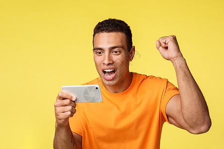 身穿橙色 T 恤 欢快兴奋的年轻帅哥举起拳头 为最喜欢的球队加油 水平拿着智能手机 在移动设备上看足球 黄色背景胜利公司成人展示图片