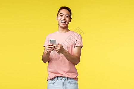 技术 通信和生活方式概念 穿着粉色 T 恤的热情亚洲男性从冷笑话或视频中大笑 使用手机 微笑着高兴的相机 站立黄色背景职业旅行情图片