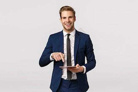 你得看看这个 穿着经典西装 领带 手持数字平板电脑和指向小工具屏幕的快乐优雅的男性企业家向商业伙伴展示在线杂志上写的精彩新闻图片