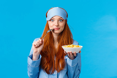 早晨 生活方式和女性观念 漂亮可爱的红头发 长着红头发的狡猾女孩早起 准备早餐 咬勺子 微笑着吃麦片 站在蓝色背景图片