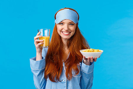 健康 食品和饮食概念 穿着睡衣 睡面罩 拿着麦片和橙汁 微笑 醒来准备早餐 站在蓝色背景中的漂亮魅力狡猾的少女图片