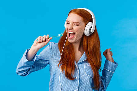 像没人看见一样唱歌 穿着睡衣的无忧无虑 兴奋快乐的红发女性 戴着大耳机 用牙刷唱歌 闭上眼睛享受美妙的音乐 站在蓝色背景中图片