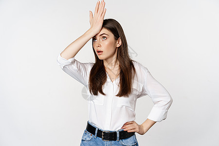 身穿白衬衫和牛仔裤 站在白底背上站着的年轻女性脸棕 拍打额头和滚动眼睛图片