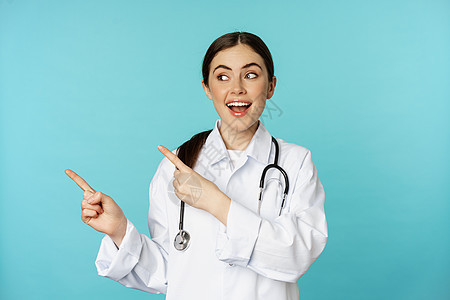 微笑的医务工作者的画像 穿白大衣的女医生拿着听诊器 手指向左 展示医疗诊所广告 绿松石背景成人擦洗临床黑发蓝色促销护士医师女士实图片
