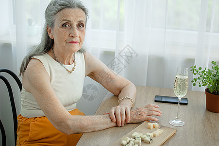 美丽的老祖母 头发灰白 脸上满是皱纹 坐在家里的桌子旁 窗户背景是一杯香槟 母亲节 退休快乐庆典饮料木板酒精舒适有钱人酒瘾幸福女图片