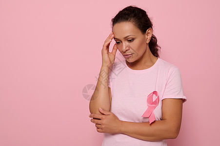 美丽体贴的女人穿着粉红色 T 恤 系着粉红色丝带 俯视着粉红色的背景 手放在太阳穴上 国际世界乳腺癌宣传日 教育 医学概念 复制图片