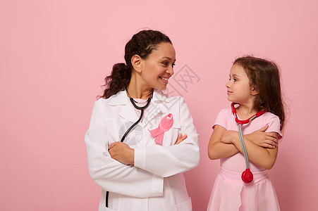 微笑的女医生和可爱的小女孩看着对方 戴着粉色丝带 这是乳腺癌宣传日的象征 双臂交叉站在胸前 彩色背景上有复制空间图片