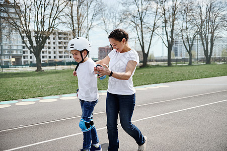 妈妈戴着头盔和防护装备站在她儿子身后 帮他在街上玩滑板游戏 母亲还帮助他图片