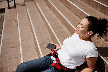 坐在台阶上的年轻美女 手里握着智能手机 在耳机上收听音乐的歌声图片