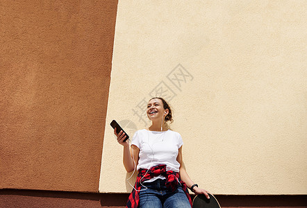 迷人的 笑着微笑的黑发女人靠在彩色墙壁上 手拿着滑板和移动电话图片