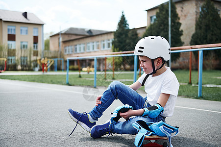 坐在滑板上并贴上防护膝盖垫的身着安全头盔的运动男孩的侧面景象图片