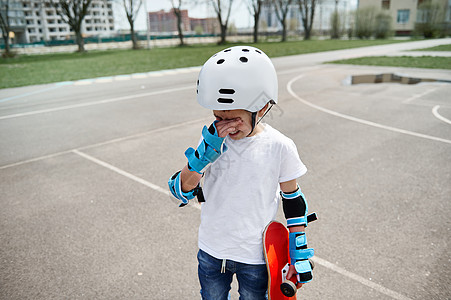 站在运动场上 穿着防护装备的心烦意乱的男孩滑板手沮丧地擦去眼中的泪水 一只手拿着滑板图片