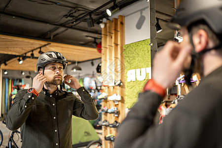 男人在运动用品商店里检查自行车头盔 看着镜子里的自己 在自行车店试新运动头盔 男性在商店里戴上自行车头盔 自行车商店的顾客试戴自图片