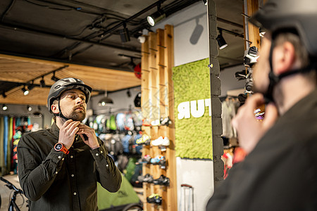 白人男子在体育用品店的镜子附近试戴黑色自行车头盔 男性买家选择骑自行车的安全头盔 在自行车商店购物 人调整他的运动头盔带零售店铺图片