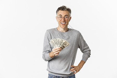 佩戴眼镜和灰色毛衣的中年人令人惊异的肖像 拿着钱 笑着欢笑 中彩票 站在白背景上中年贷款胡须现金投资促销发型商业情感广告图片