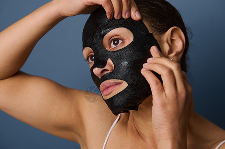 美丽的女人在脸上戴上保湿平滑滋养清洁黑色织物面膜 隔离在灰色背景中 有复制空间 护肤 美容 家庭水疗概念 特写图片