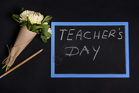 平铺的木铅笔和一束美丽的紫苑花 用工艺包装纸放在粉笔板旁边 上面写着教师节的字样 在黑色背景中被隔离 有复制空间图片
