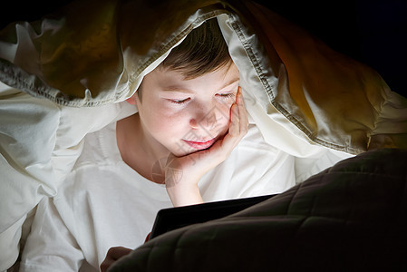 远程教育 在线教育 快乐的男孩躺在床上 毯子下 黑暗中有平板电脑 明亮的监视器照亮了孩子的脸 隔离 社交媒体成瘾阅读视频儿童互联图片