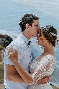 美丽的新娘和新郎在希腊岛圣托里尼的夏季婚礼当天 在婚纱下拥抱和亲吻蜜月晴天夫妻妻子玫瑰庆典女士花朵天空家庭图片