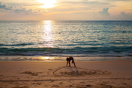 日落时 女孩在靠近海的沙滩上将心涂在沙子上图片