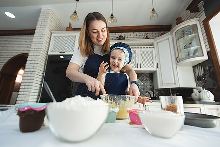 母亲和女儿穿着相同的围裙和厨师帽在厨房里做饭 他们用木铲搅拌面团 微笑着看着镜头图片