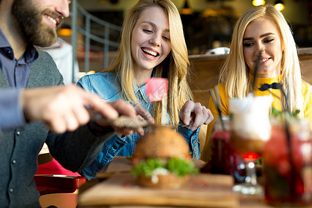 快乐的朋友们在餐馆里用刀砍了汉堡包 然后笑派对友谊酒吧女孩桌子小吃蜜饯食物芝士咖啡店图片