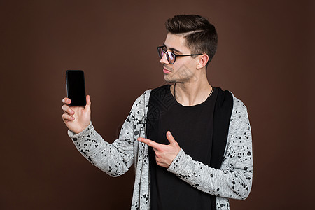 年轻英俊男性模特的近距离肖像 把智能手机对着摄影机 与棕色背景隔绝黑色商业工具屏幕展示细胞空白成人互联网技术图片