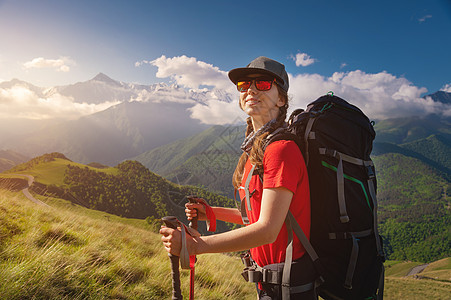 在高山和低云的背景下 戴着帽子和太阳镜的白人旅行者背着背包和登山杖 微笑着看着相机图片