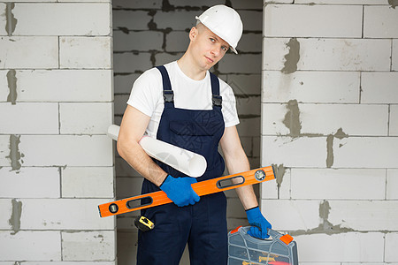 男性工人或建筑工人戴着头盔 头盔上有水平仪 工具箱和蓝图 贴在砖墙上图片