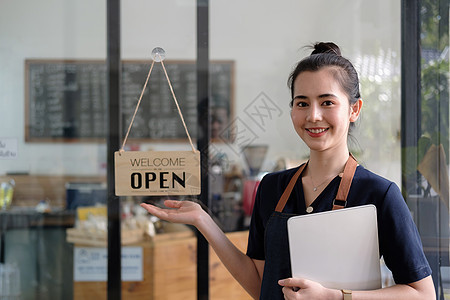 美丽的亚洲年轻咖啡师女人穿着围裙拿着平板电脑 站在咖啡馆门前 上面有开放的招牌 企业主创业中小企业企业家的概念图片