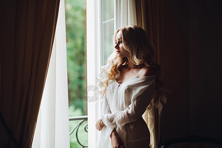 美丽的新娘站在阳台旁边 看着大窗的风景白色内衣身材金发卷曲婚礼女性窗户化妆品房间图片