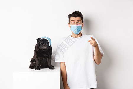 Covid19 动物和检疫概念 快乐的狗主人和戴着医用口罩的可爱哈巴狗 男人用手指指着镜头惊讶 白色背景微笑小狗潮人行动促销广告图片