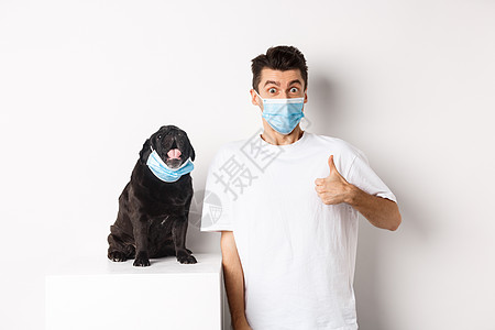 Covid19 动物和检疫概念 有趣的年轻人和小狗在医疗面具中的形象 主人举起拇指表示认可 赞美某些东西 白种背景宠物乐趣促销广图片
