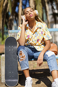 坐在滑板上听音乐的非洲女孩在骑滑板后放松了一下发型耳机音乐微笑女性溜冰者辫子恐惧青年闲暇图片