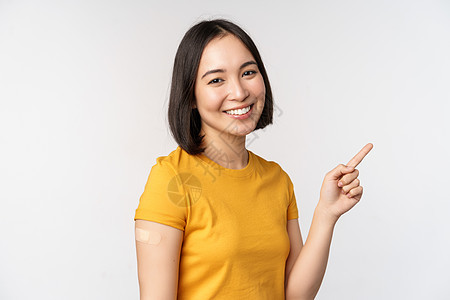 Covid19 和疫苗接种概念 微笑的亚洲女孩戴着肩带 用手指指着横幅 展示疫苗运动 站在白色背景上广告成人工作室大学情感女性商图片