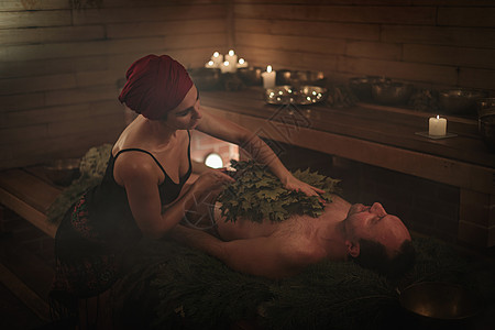 一名妇女在扫帚的帮助下为一名男子举行沐浴仪式 一个人躺在云杉树枝上疗法治愈者享受乐趣成人蜡烛治疗蒸汽桑拿精神图片