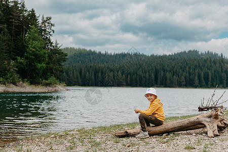 小男孩坐在山湖附近的圆木上 一个孤独的男孩静静地坐着 看着前方美丽的山景 小男孩明尼苏达湖 小男孩湖度假村假期旅行蓝色男性孩子们图片