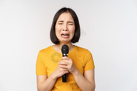 站在白色背景上 哭泣的亚洲女孩在麦克风里唱着伤心欲绝的歌 拿着麦克风 心烦意乱地做着鬼脸女朋友成人情感唱歌大学情绪压痛互联网工作背景图片